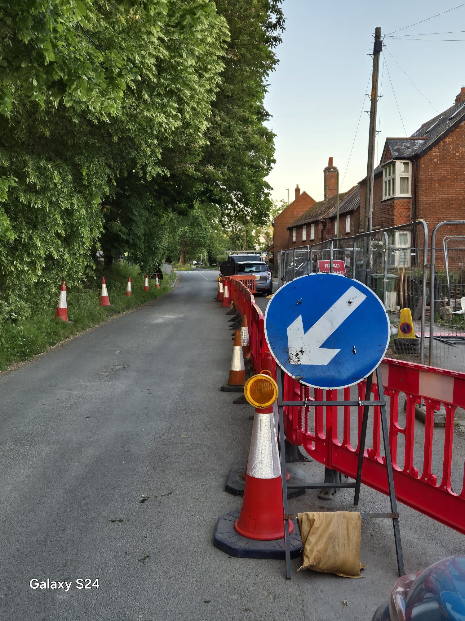 Keep left arrow sign, cones and red barriers narrowing a village road past a fenced works compound