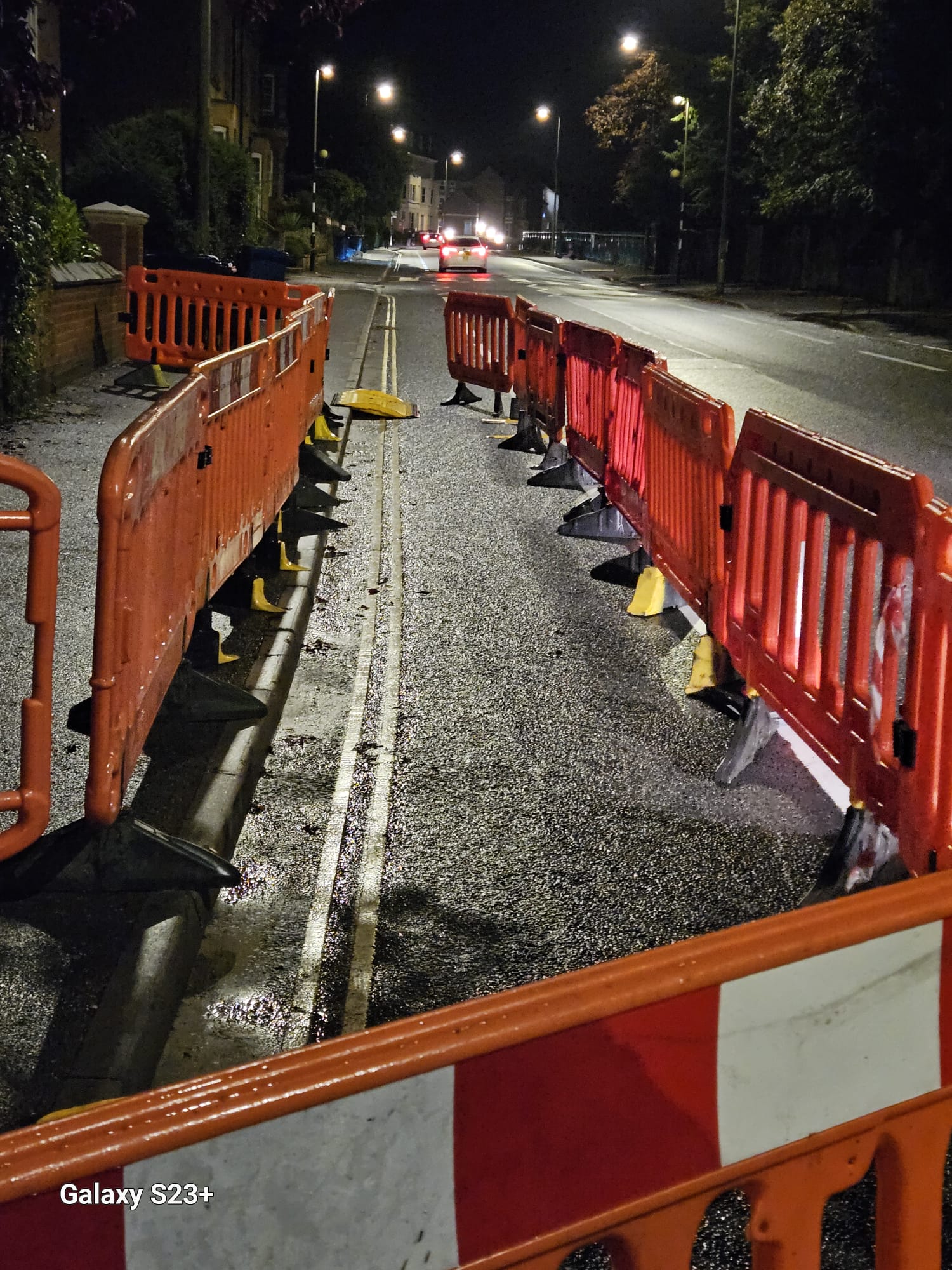 Barriered lane closure on a wet residential road at night with traffic passing the works area
