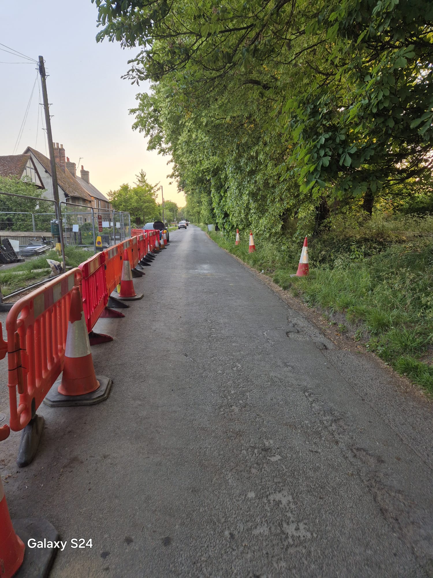 Long barrier and cone run holding a single-lane working space along a village road at dusk