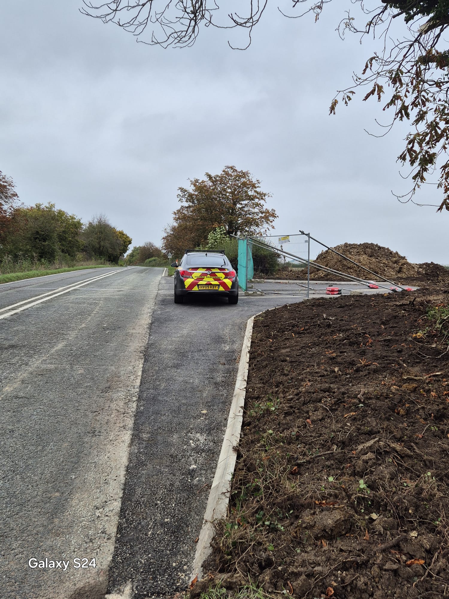 Highway maintenance vehicle parked at a fenced rural site access alongside freshly laid kerbs and surfacing
