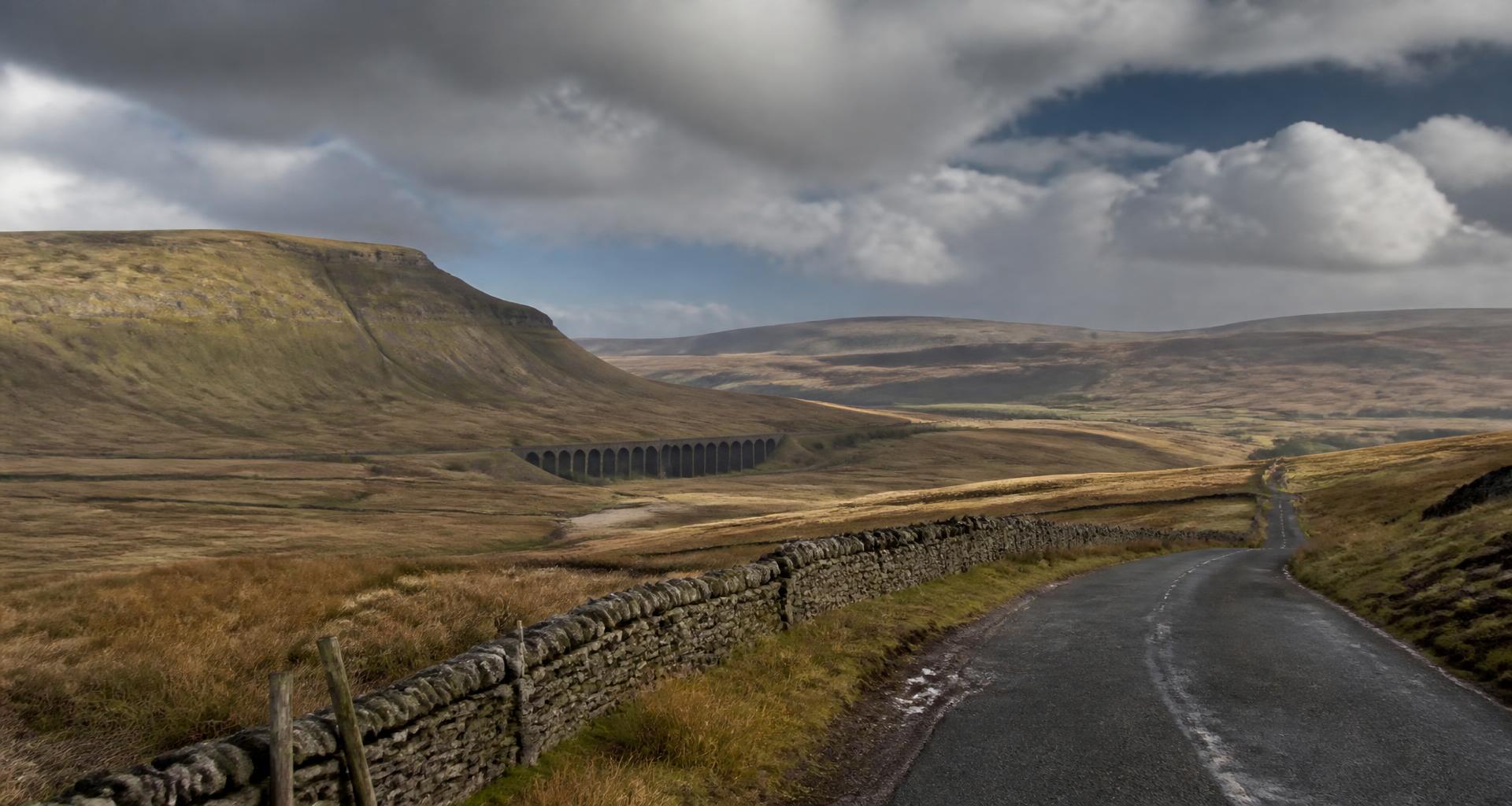 Northern England road climbing across Pennine moorland with dry-stone walls and a stone viaduct
