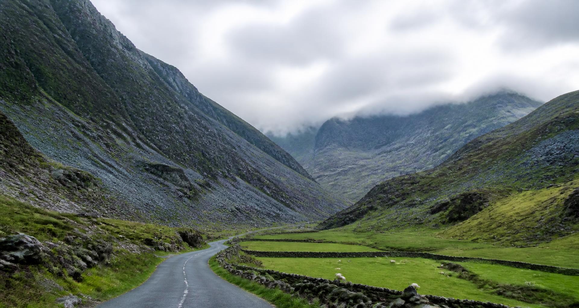 Narrow Welsh mountain road winding through a green valley beneath slate-grey rock faces